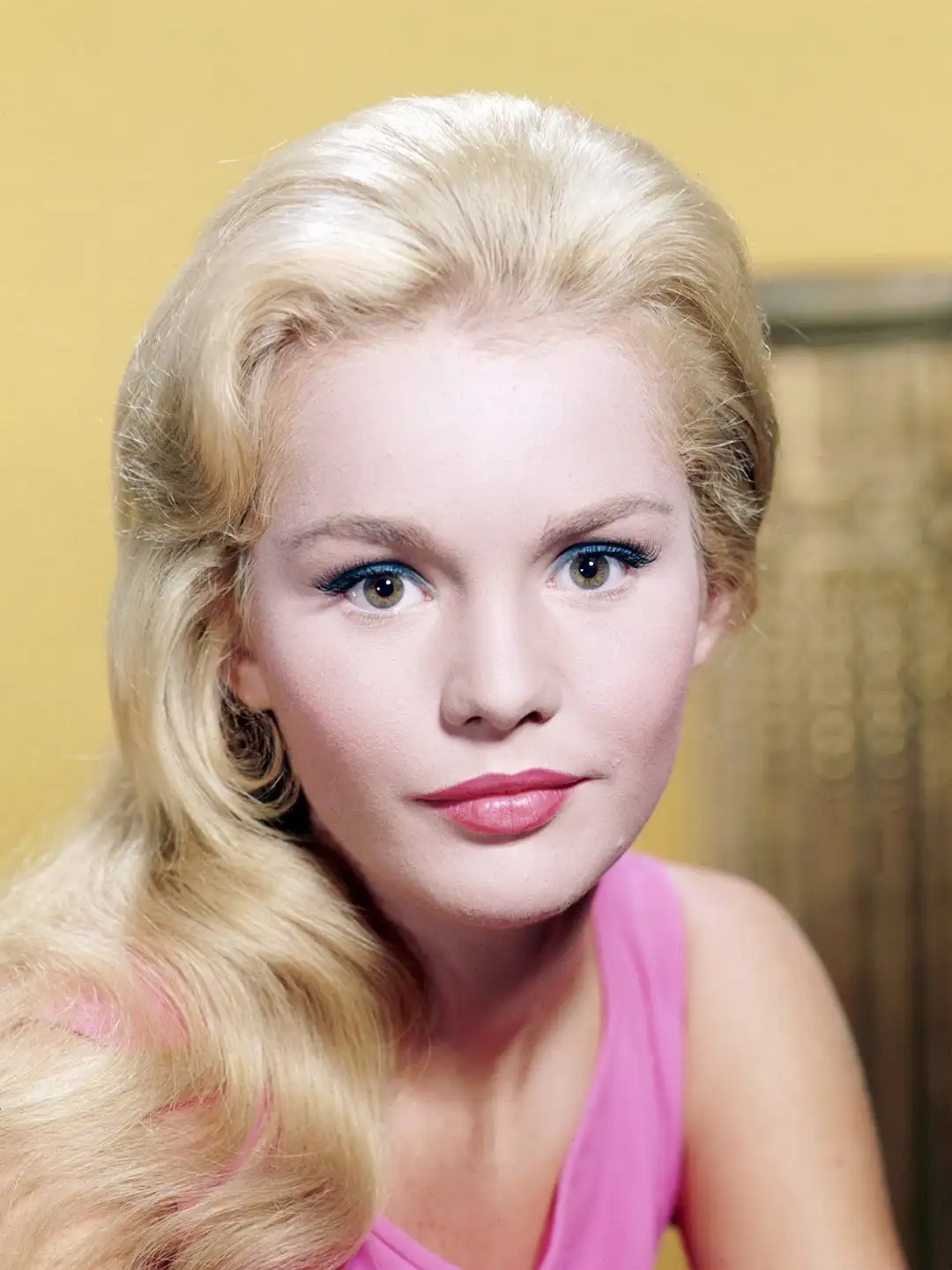 A vintage studio portrait of a blonde woman with voluminous hair, wearing a pink top and striking blue eyeliner, looking…