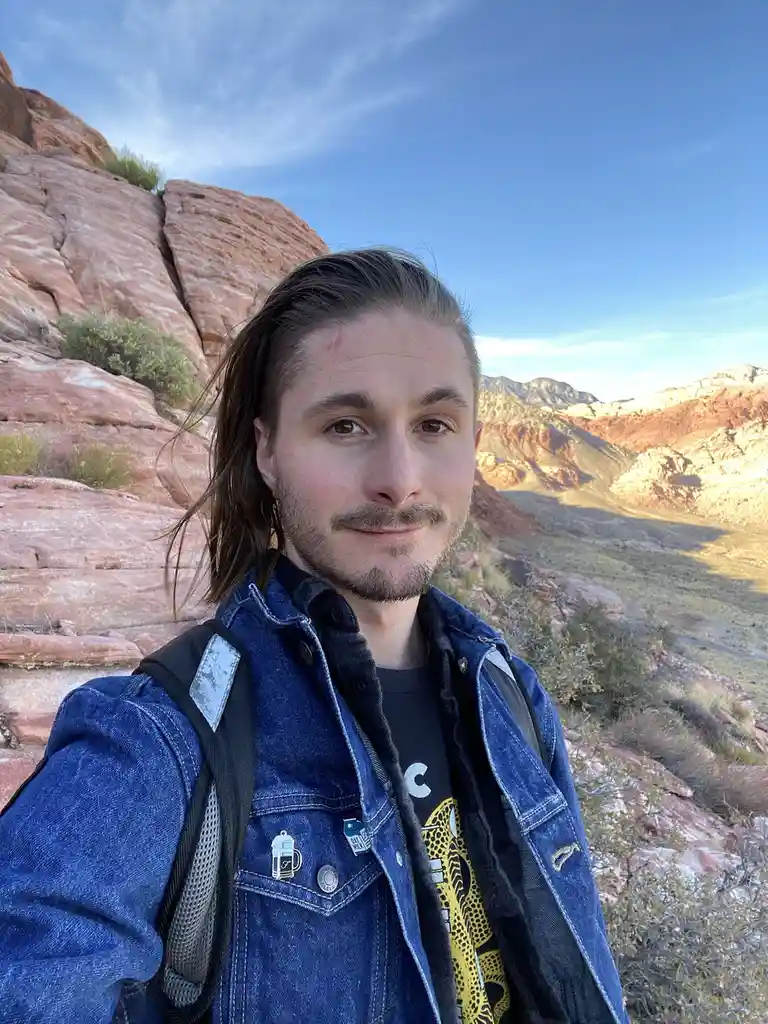 A man with long hair and a beard, wearing a denim jacket over a graphic tee and carrying a backpack, takes a selfie against…