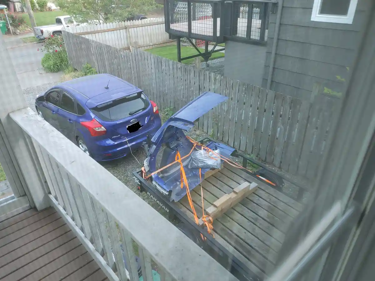 A blue Ford Focus is parked in a gravel driveway next to a trailer. The trunk of the car is open and the rear body panel is…