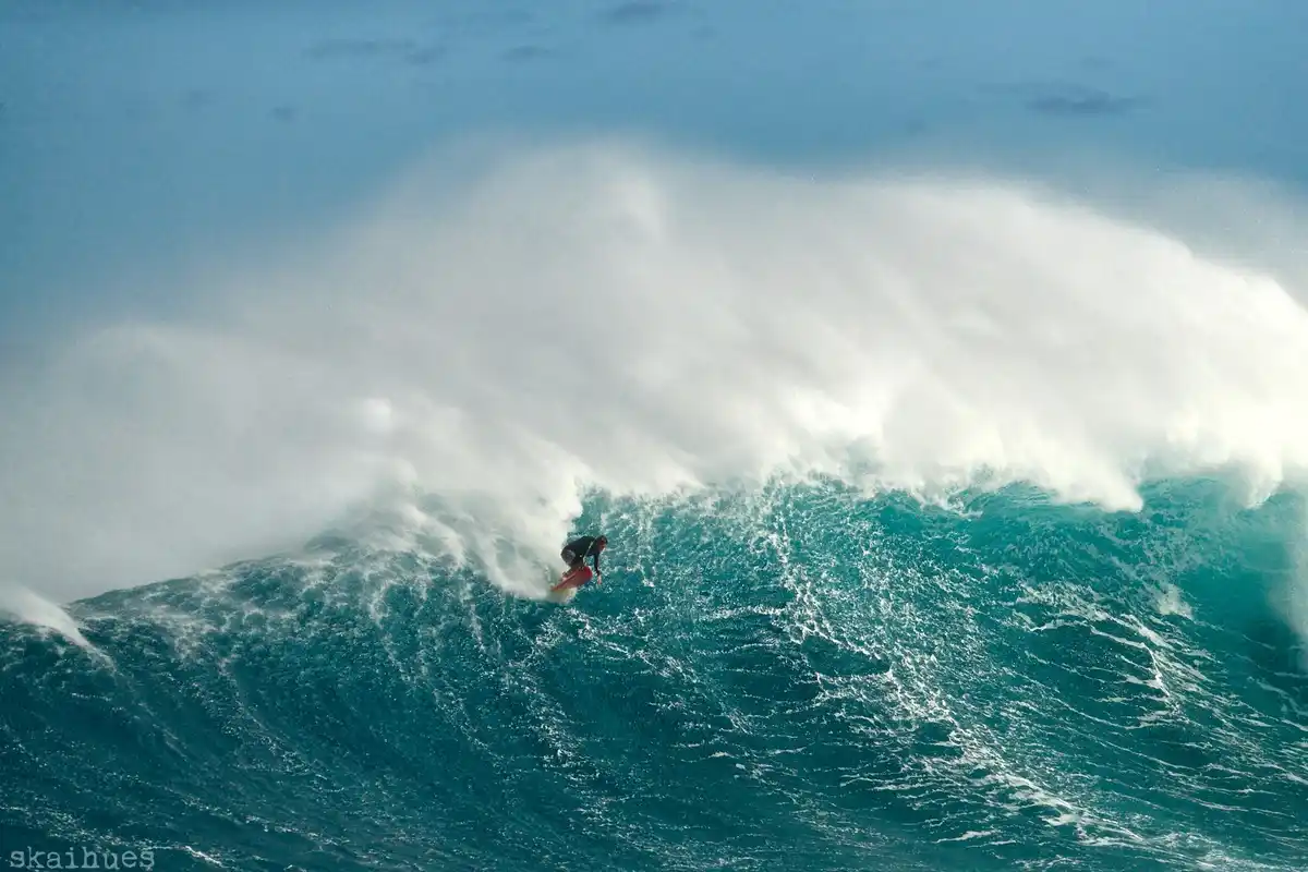 A surfer in a black wetsuit rides a massive, turquoise ocean wave with a towering wall of white spray behind them. The…