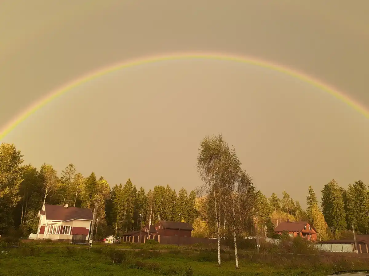 A wide-angle photo shows a full, vivid rainbow arcing across a hazy, golden sky above a rural landscape with scattered…