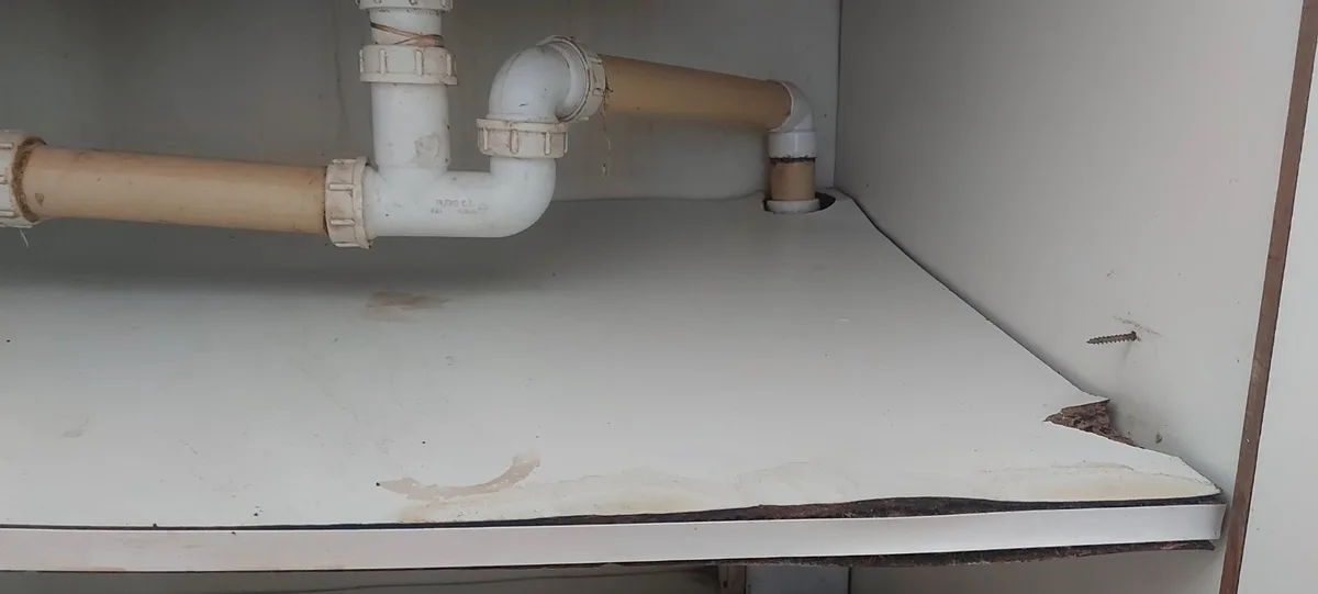 A photo shows the underside of a white cabinet with visible plumbing pipes, a broken corner, and water stains. The overlaid…