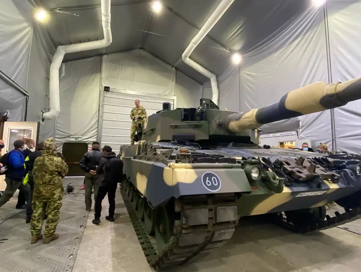 A large military tank with the number 60 painted on its front is displayed inside a large tent. A soldier in camouflage…