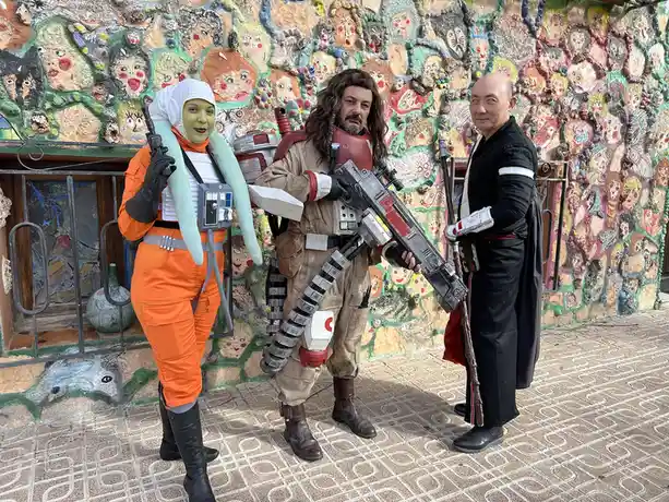 Three people dressed as Star Wars characters — a green-skinned alien in orange jumpsuit, a bounty hunter with a large…