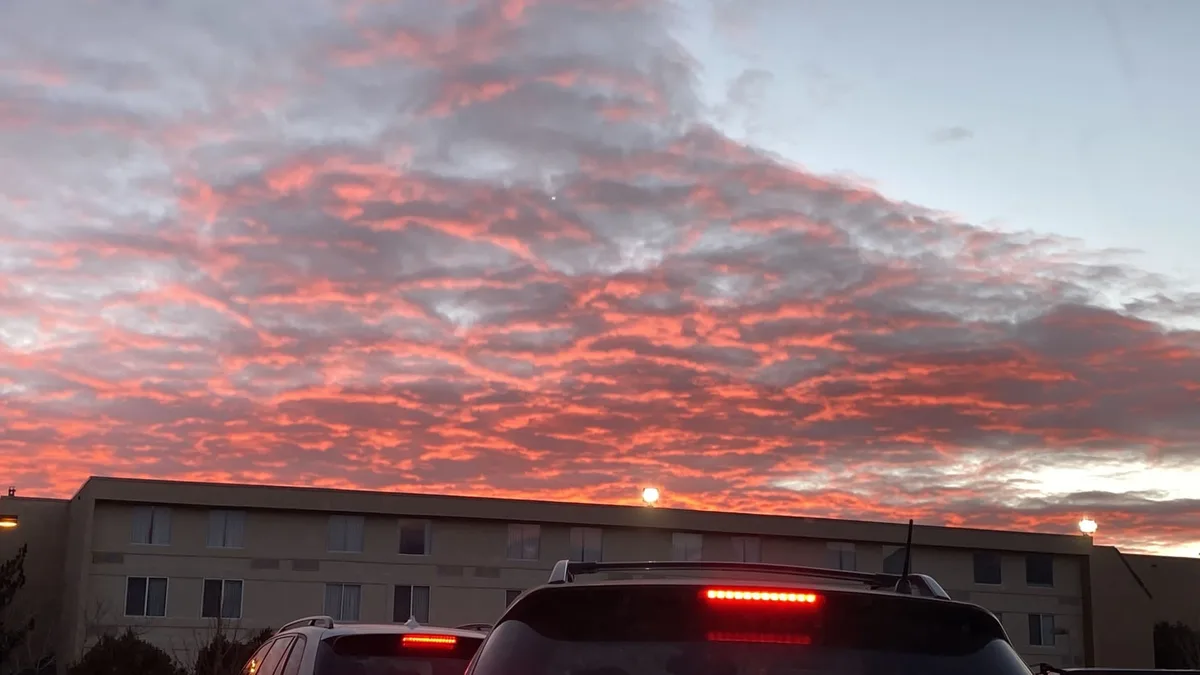 A photo of a vibrant, fiery orange and pink sunset or sunrise over a multi-story apartment building with cars parked in…