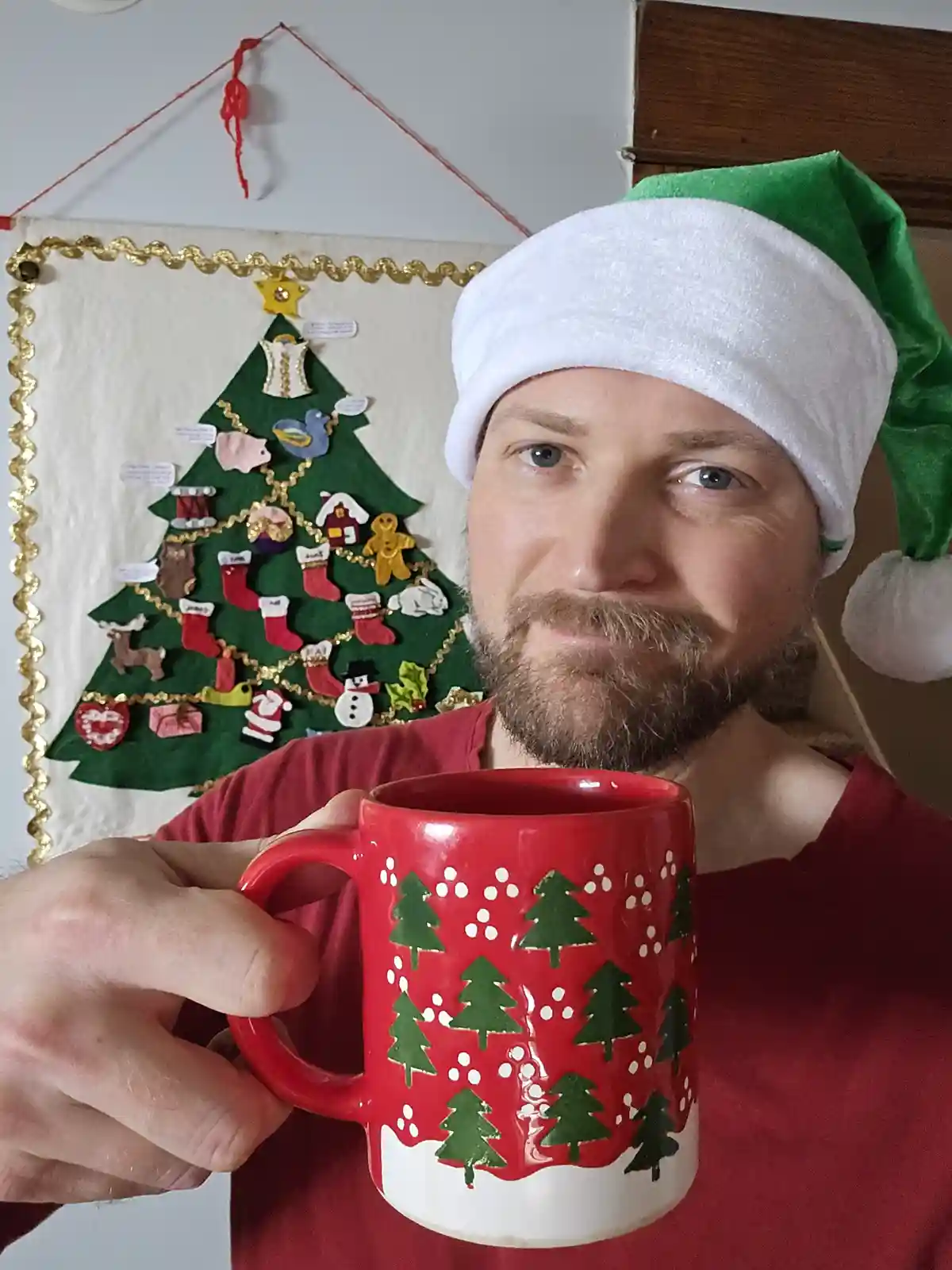 A man with a beard wearing a green and white Santa hat and a red shirt holds a red mug with green Christmas trees and white…