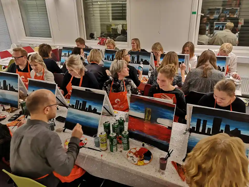 A group of people in orange aprons sit at a long table, each painting a cityscape on canvas. Most are painting a similar…