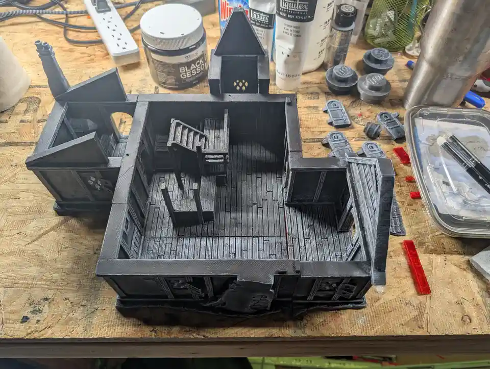 A 3D-printed miniature house model, mostly painted black, sits on a wooden workbench surrounded by art supplies like paint…