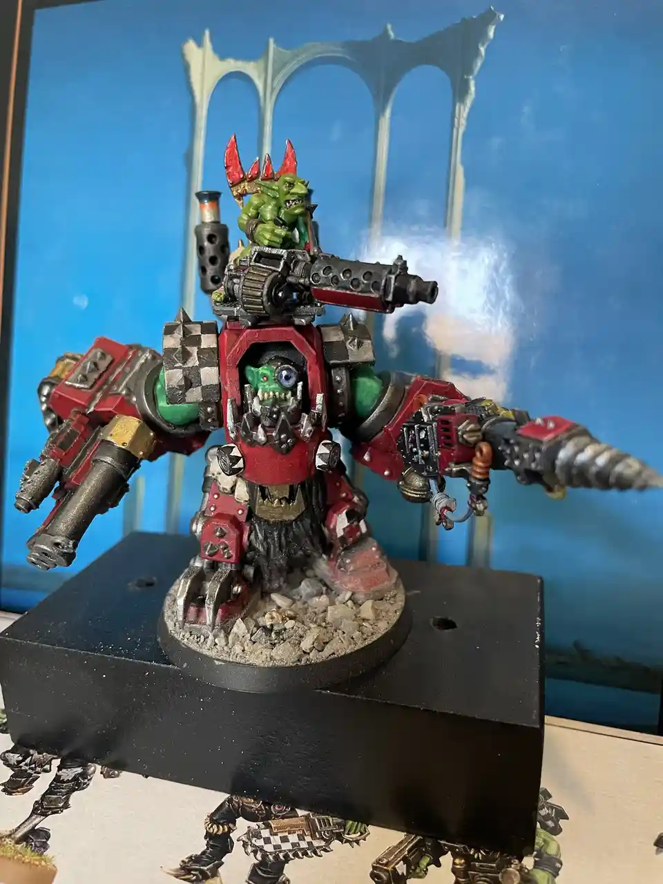 A heavily armored, red-and-green painted goblin mech with multiple weapons and a goblin pilot on top, standing on a rocky…