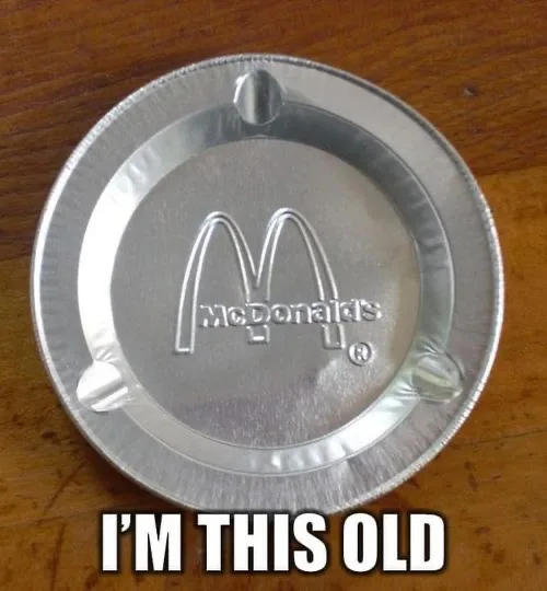 A close-up photo of a silver McDonald's branded pie tin resting on a wooden surface. The tin features the iconic golden…