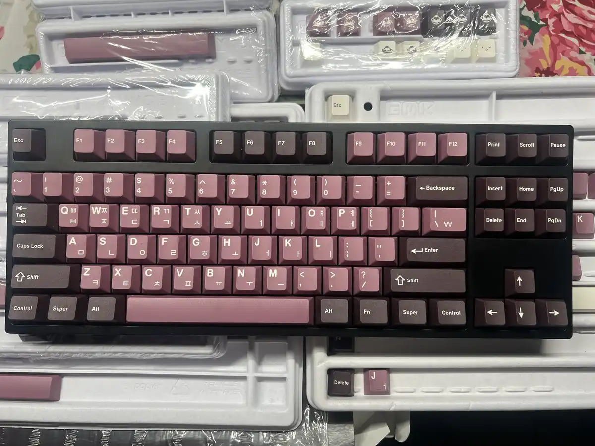 A close-up photo shows a black mechanical keyboard with pinkish-brown keycaps, resting on its white plastic packaging…