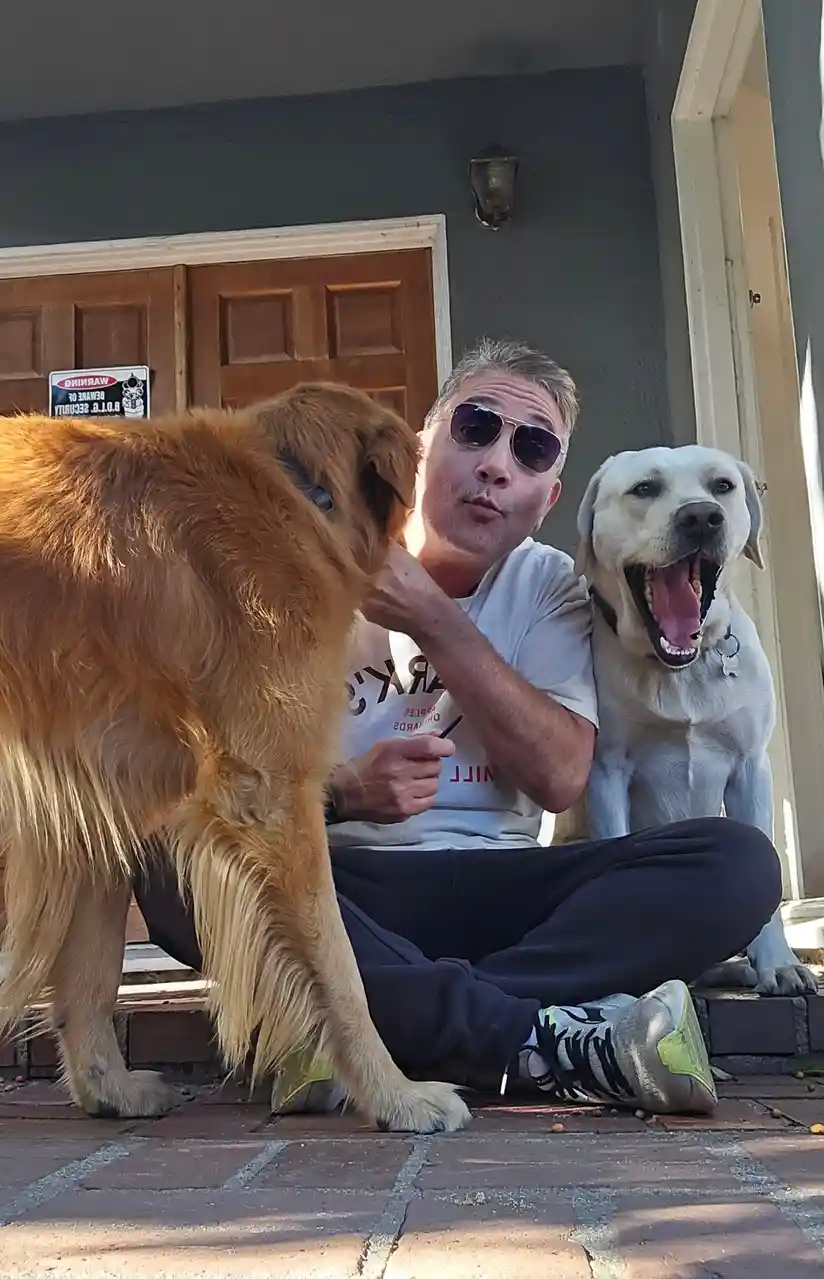 A man wearing sunglasses sits on a porch step with two dogs, one golden retriever and one yellow lab. He makes a playful…