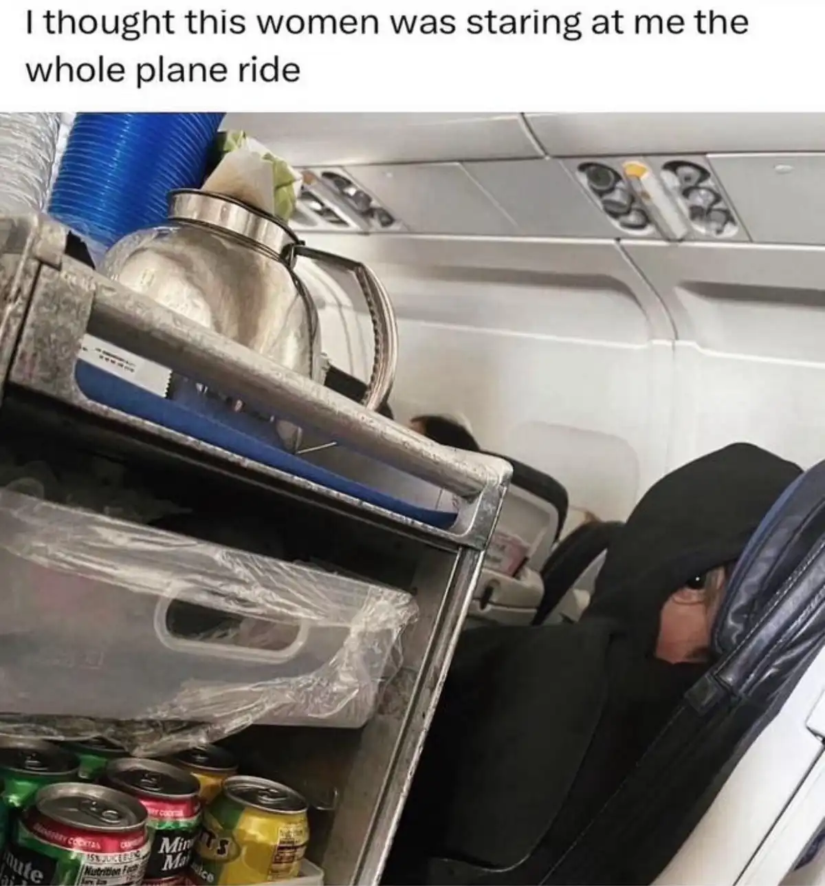 A person wearing a black hoodie sits in an airplane seat, peeking out from under the hood with only one eye visible. In…