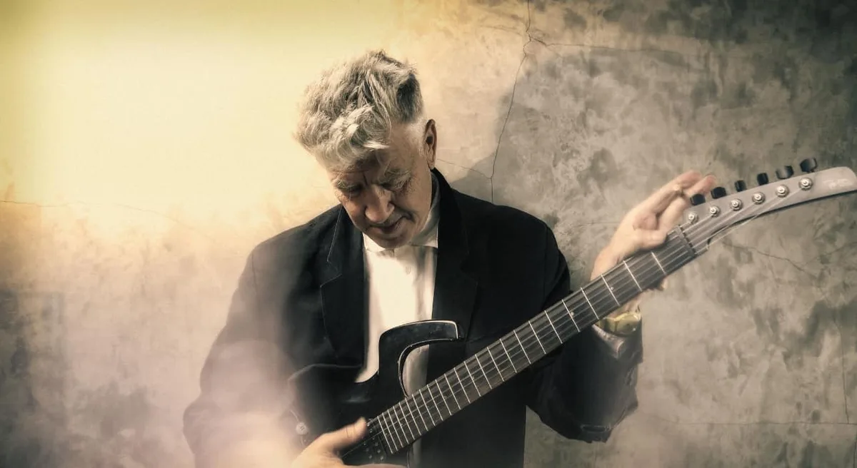 An older man with gray hair, wearing a black suit and white shirt, is playing a black electric guitar against a cracked…
