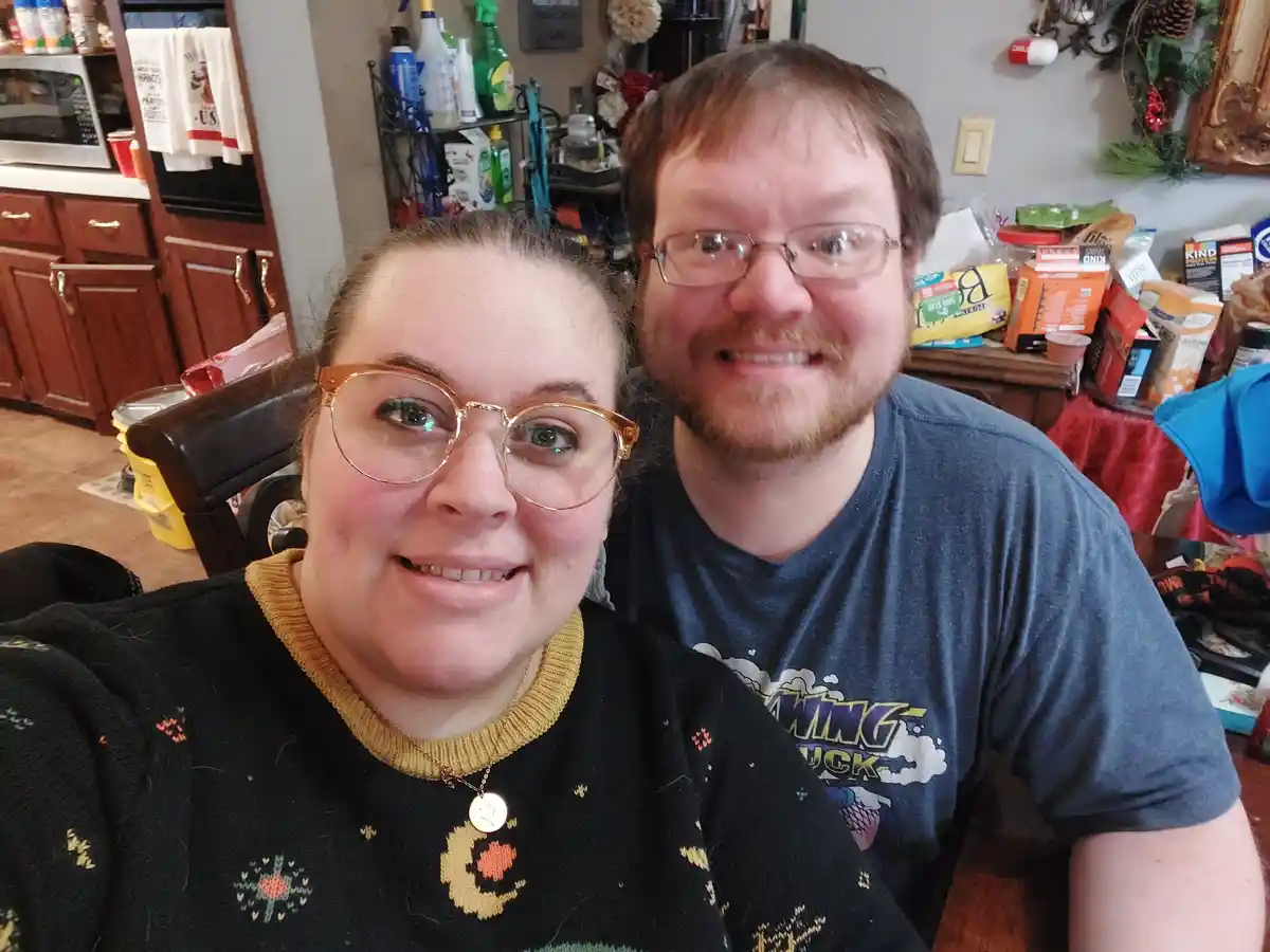 A selfie of a smiling woman in a patterned black sweater and gold-rimmed glasses and a man in a blue t-shirt with glasses…