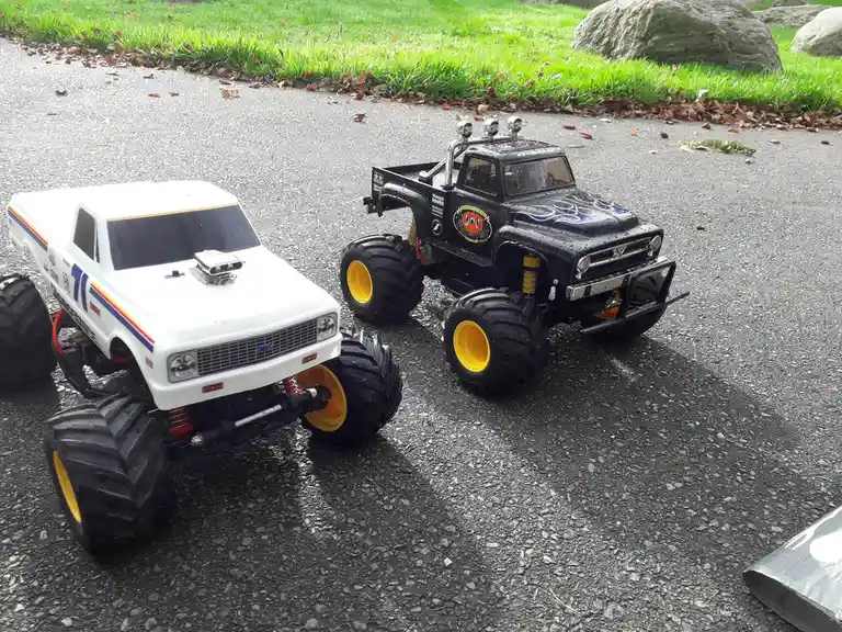 Two remote-controlled monster trucks, one white and one black with flame decals, sit on a wet asphalt driveway. The white…