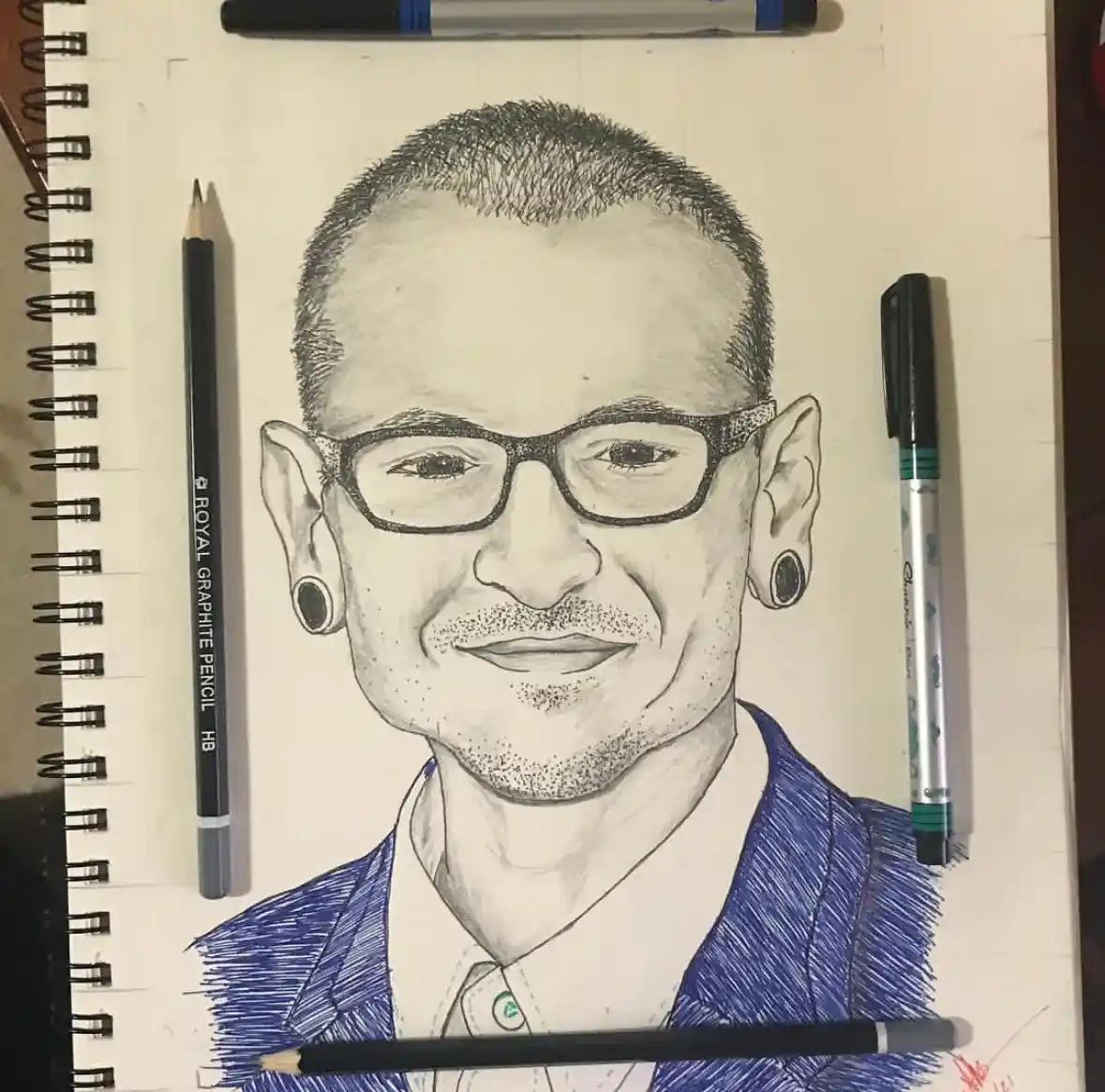 A detailed pencil and marker portrait of a smiling man with glasses and ear gauges, drawn on spiral-bound paper, with…