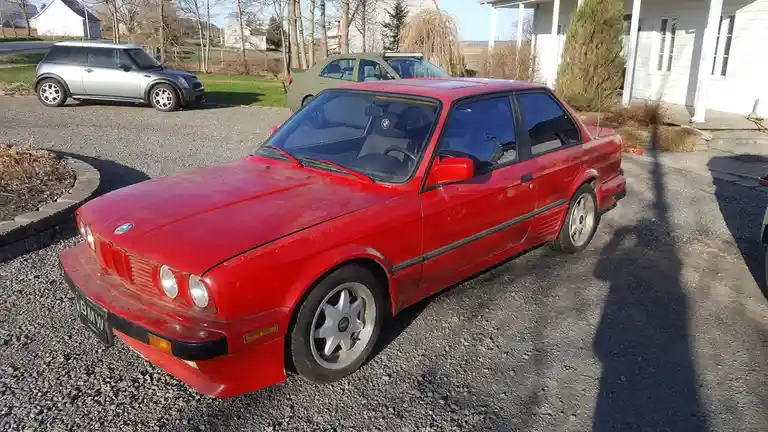 A red BMW E30 coupe is parked on a gravel driveway in front of a white house, with a silver Mini Cooper and a green sedan…