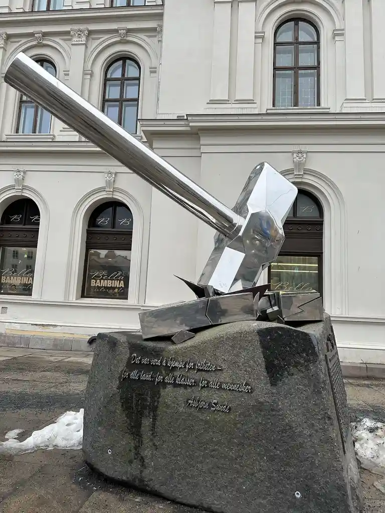 A large, shiny, metallic sculpture of a hammer and sickle, appearing to be broken or shattered, sits atop a dark stone…
