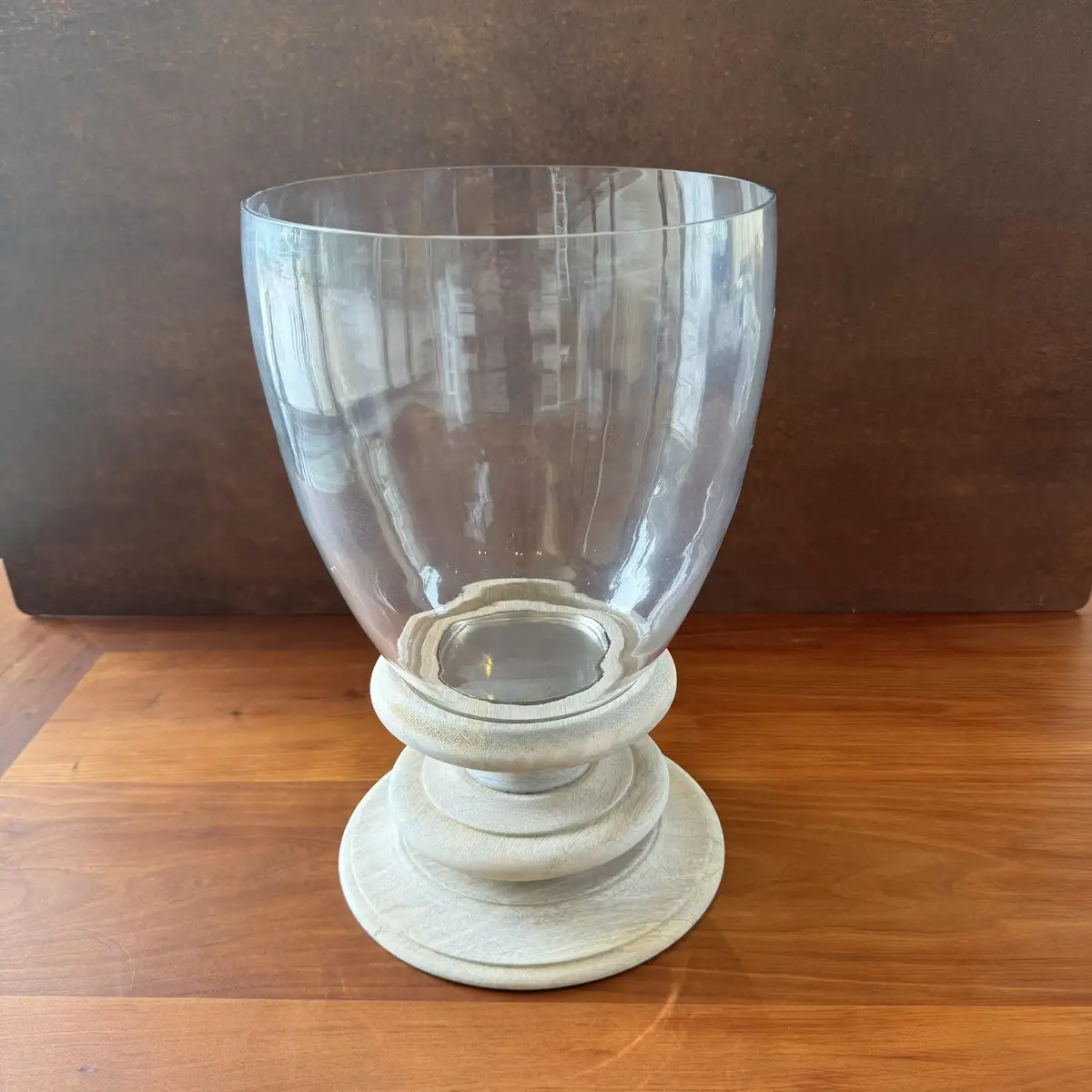 A clear glass vase with a flared top sits on a white wooden pedestal base, placed on a wooden table against a brown wall…