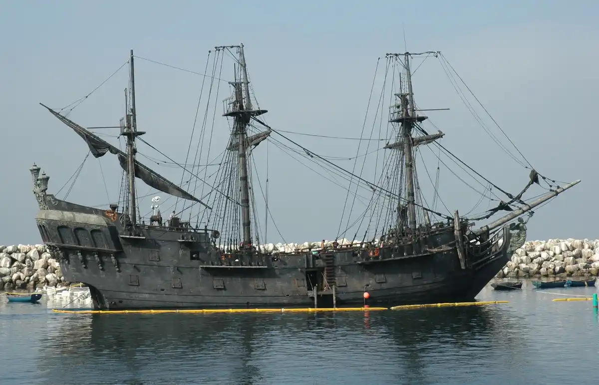 A large, dark-hulled, three-masted pirate ship with tattered sails is docked in calm water next to a rocky breakwater…