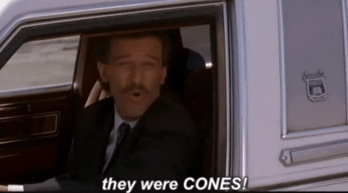 A man with a mustache in a suit sits in the driver's seat of a white hearse, looking shocked with his mouth open. The…