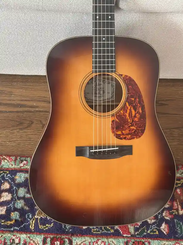 A close-up photo of a sunburst-finish acoustic guitar with a decorative pickguard, resting on a patterned rug and wooden…