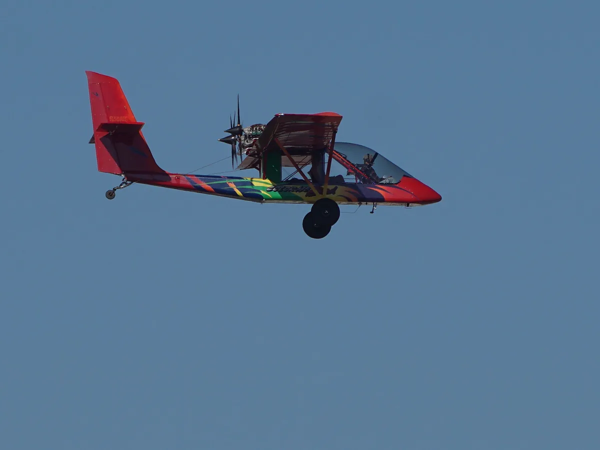 A small, colorful homemade-looking airplane with a red tail and multi-colored fuselage is flying against a clear blue sky…