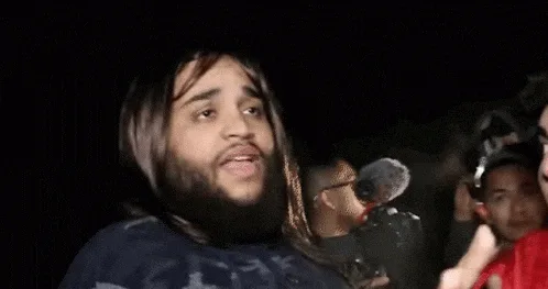 A six-panel image sequence shows a man with long hair and a beard, wearing a dark shirt, surrounded by people with cameras…