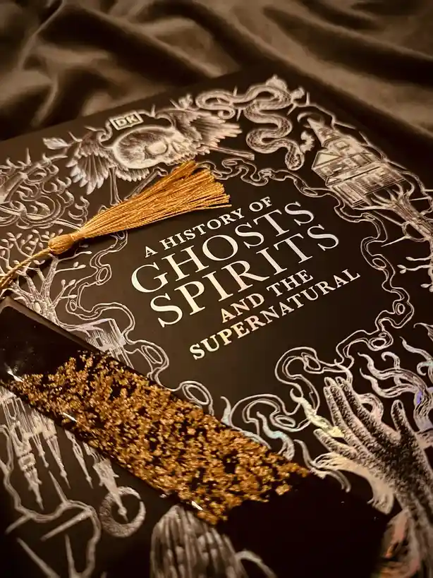 A close-up of a black book titled 'A History of Ghosts, Spirits and the Supernatural' by DK, featuring intricate white line…