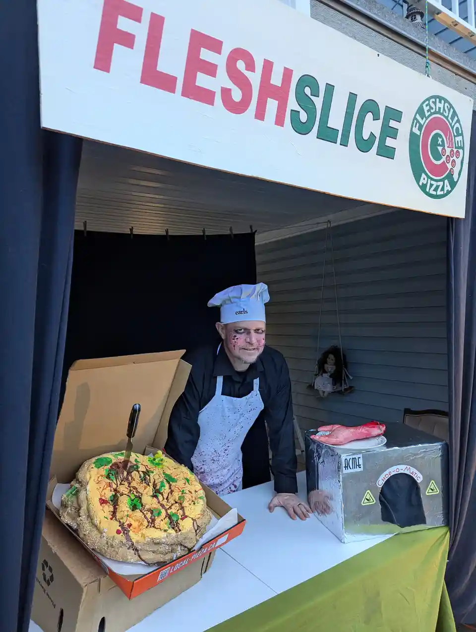A man in a chef's hat and blood-splattered apron stands behind a counter under a sign that reads 'FLESHSLICE PIZZA'. On the…