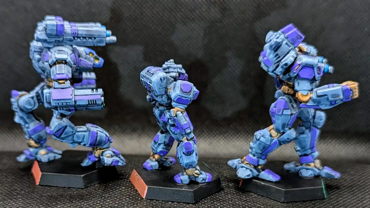 Three blue and purple painted robot miniatures stand on black bases against a dark textured background. The robot on the…