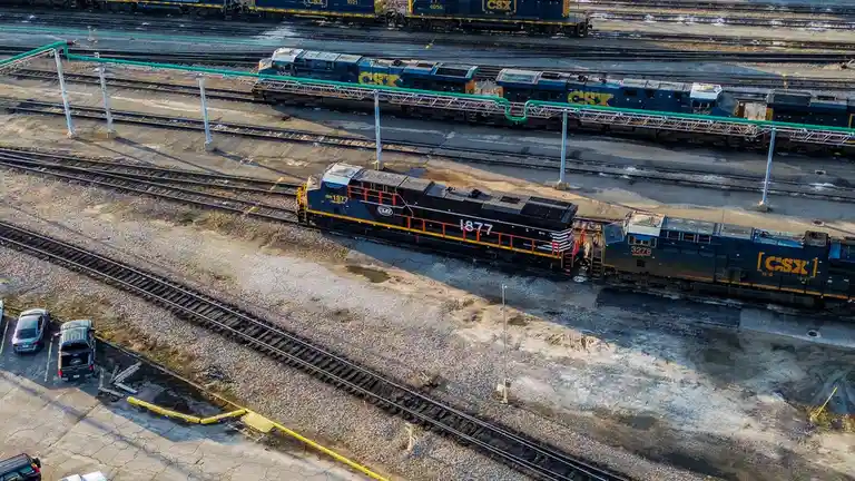 An aerial photo shows a black CSX locomotive numbered 1877, with orange railings, parked on a rail yard track next to other…