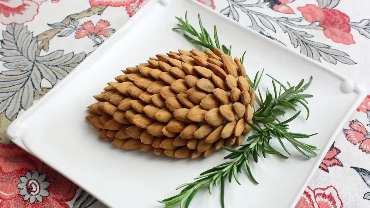 A white square plate holds a decorative arrangement resembling a pinecone, crafted from numerous almonds arranged in…