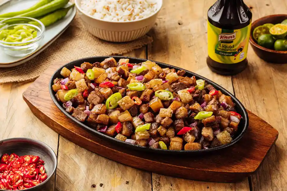 A professionally styled food photograph shows a sizzling black iron skillet filled with crispy, caramelized pork belly…