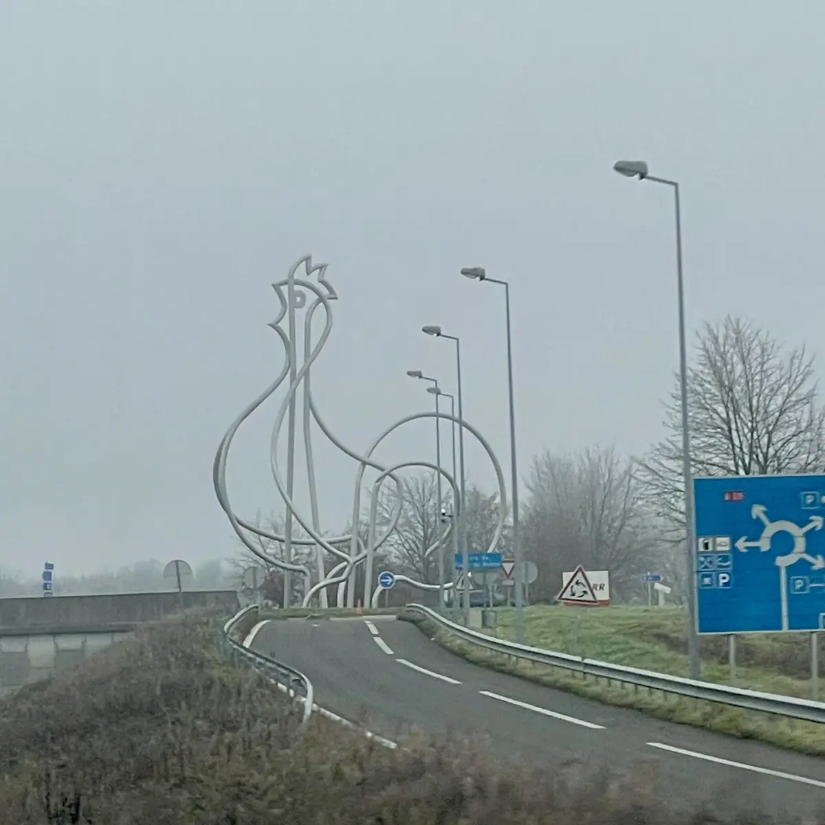 A large, abstract metal sculpture of a chicken stands beside a road under an overcast sky, with streetlights and a blue…