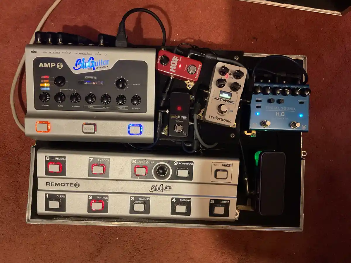 An overhead shot of a densely packed guitar pedalboard inside a metal case, featuring a BluGuitar Amp 1 unit, a red HOF…