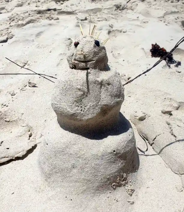 A sand sculpture shaped like a snowman stands on a sunny beach, made of three stacked rounded sections. It has twig arms…