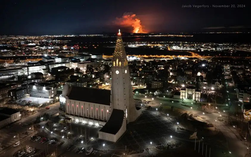a nighttime aerial photo shows a brightly lit modern church in a city, with a large volcanic eruption glowing orange on the…