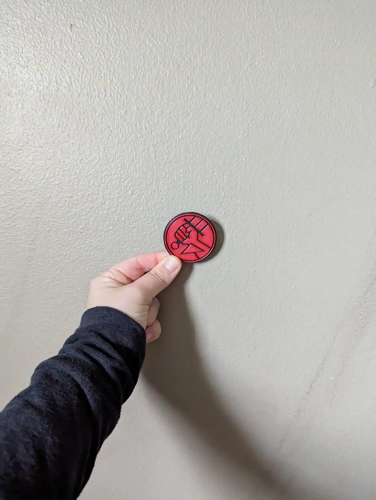 A hand holds a circular red and black pin featuring a stylized fist logo against a plain beige wall. The overlaid text reads…