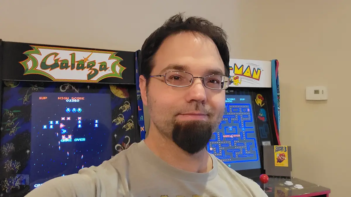 A man with glasses and a goatee takes a selfie in front of two arcade machines, both displaying 'GAME OVER'. The machine on…