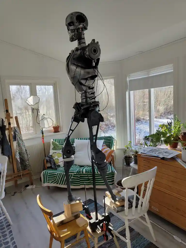 A tall, black, skeletal robot with a skull head and exposed wiring stands in a cozy, well-lit living room with white walls…
