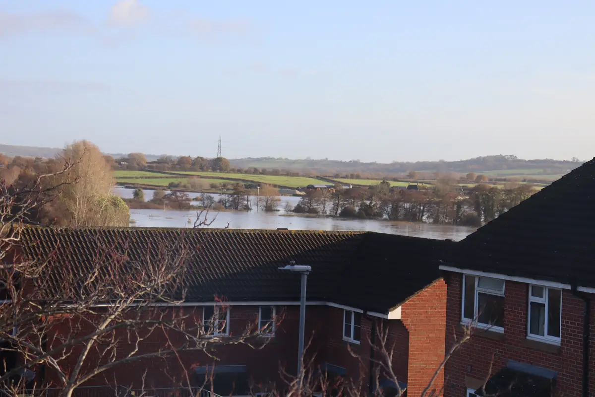 A photo shows a flooded rural landscape viewed from a window, with red brick houses in the foreground and water covering…