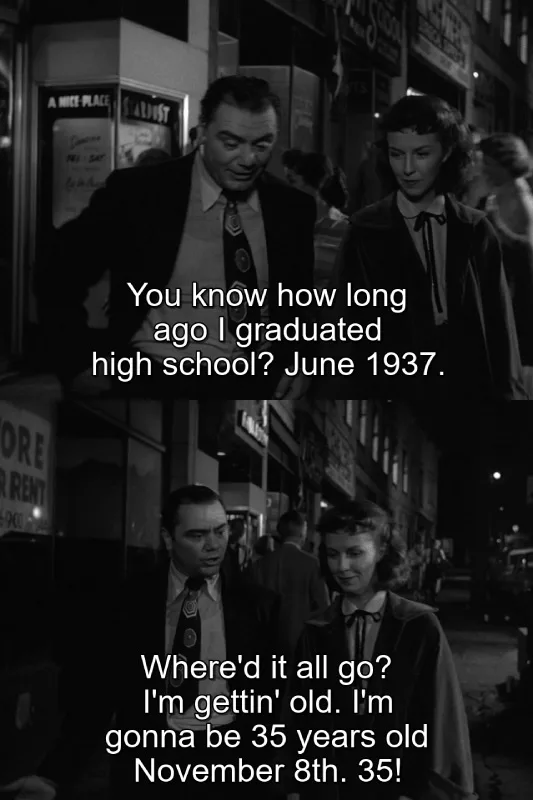 A black-and-white still from a classic film shows a man and woman walking down a city street at night. The man, looking…