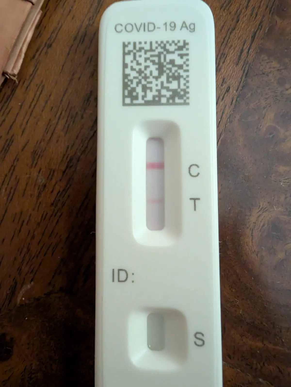 A close-up photo of a white COVID-19 antigen test strip resting on a dark wooden surface, showing two pink lines in the…