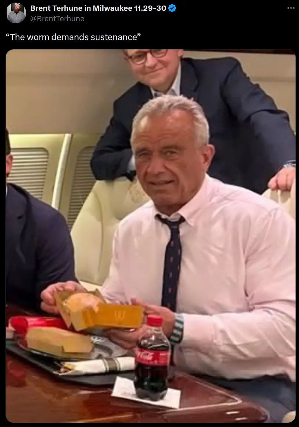 A man in a white shirt and tie sits at a table with a meal tray, holding a box of what appears to be a sandwich or bread. A…