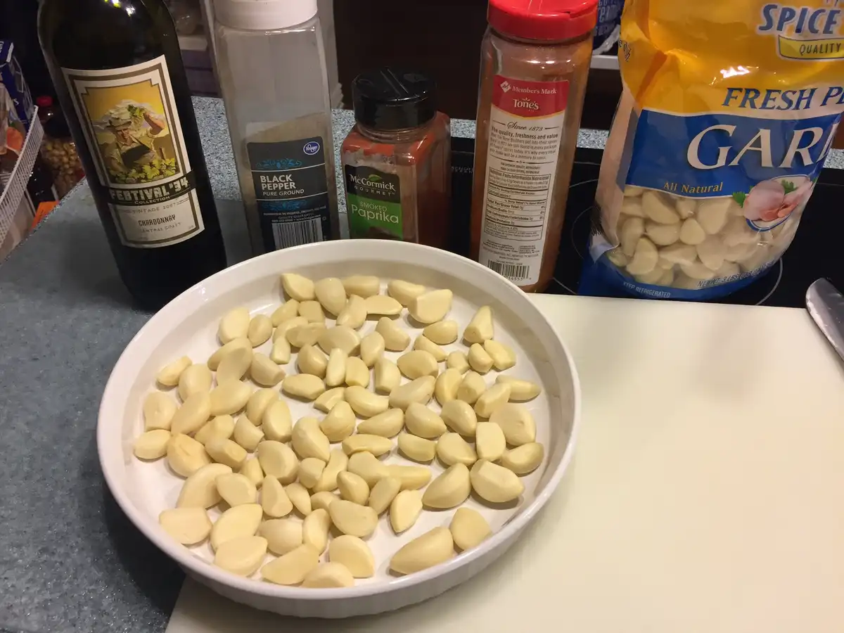 A white bowl filled with peeled garlic cloves sits on a kitchen counter, surrounded by a bottle of Chardonnay wine, a…