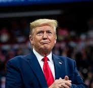 a photo of Donald Trump in a dark suit and red tie, clasping his hands together with a smug, self-satisfied expression. The…
