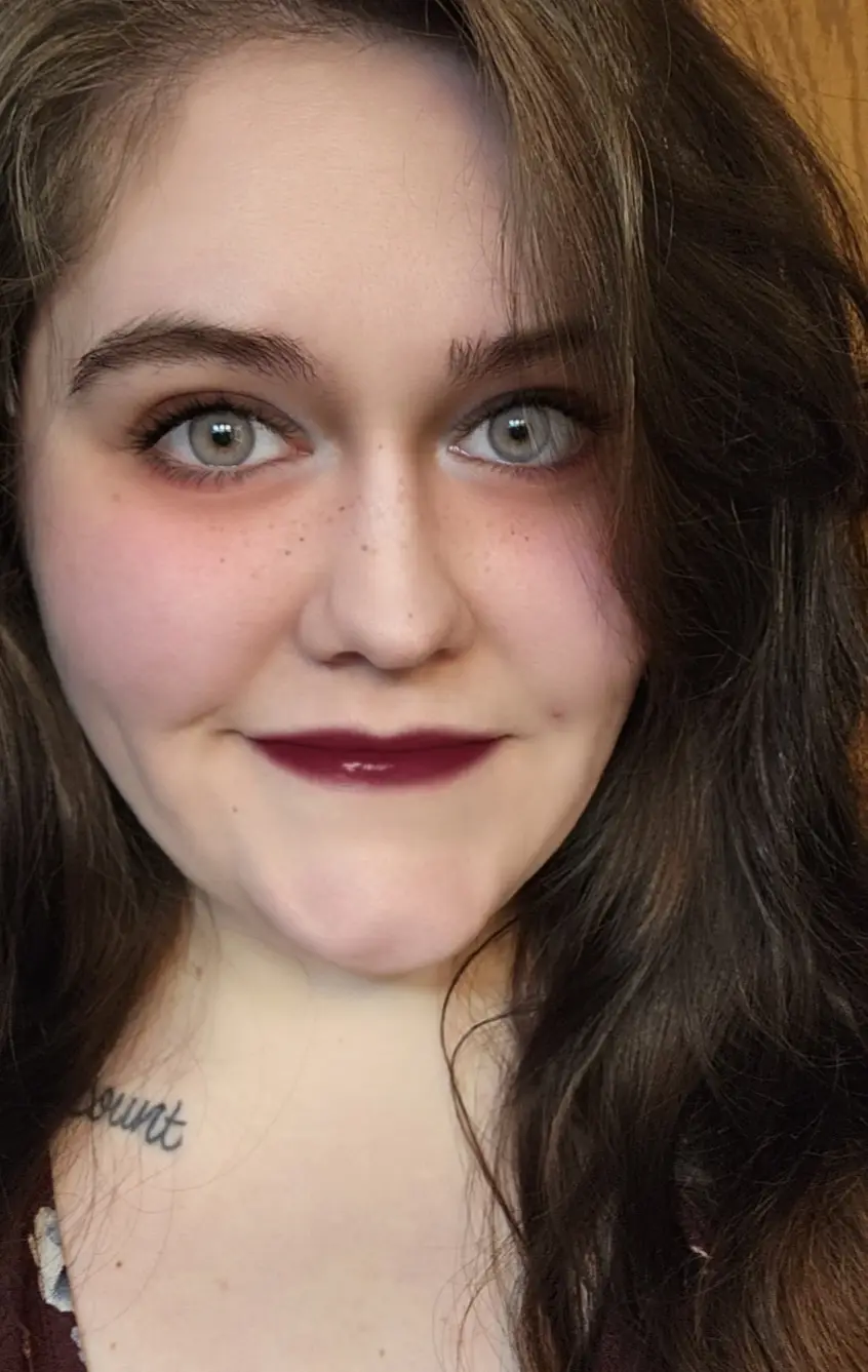A close-up selfie of a young woman with brown hair, freckles, and dark red lipstick, smiling slightly. She has a small…