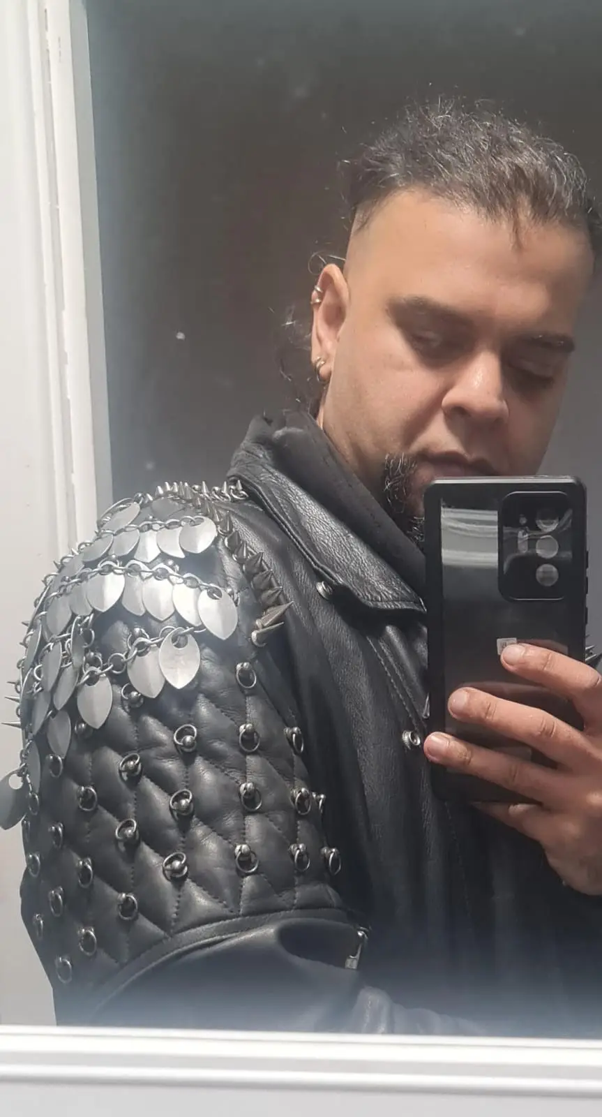 A man with a shaved head and goatee takes a mirror selfie wearing a heavily modified black leather jacket adorned with metal…